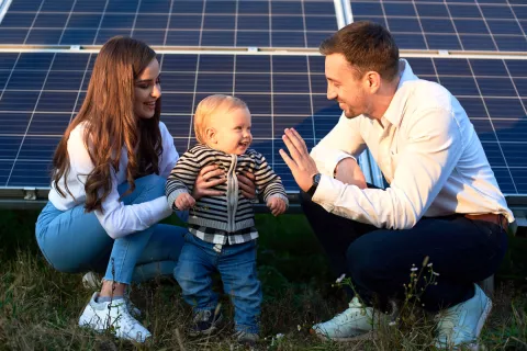 Ein junger Vater und eine junge Mutter lachen und sitzen vor einem Solarpanel in der Sonne.